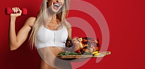 Closeup of smiling sexy fitness woman in white sport bra holding whole roasted chicken on tray and dumbbell in hand