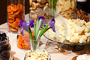 Closeup snacks, dried fruits and nuts in glass bowl, flowers irises in vase, plate with pieces with parmesan cheese. Concept