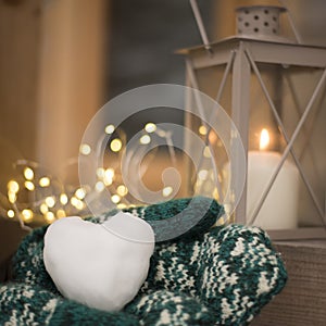 Closeup of snowball in shape of heart in hands with mittens and outdoor candle holder with burning candle