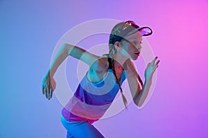 Closeup of sportive girl, junior runner in stylish sportswear and cap posing isolated on gradient pink-blue background