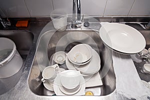 Closeup stack of dirty dishes at bottom of metal square sink at professional restaurant kitchen: white ceramic stack of plates,