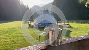 Closeup of a steaming metallic cup with hot coffee or tea in the morning with glamping camping ground in the background