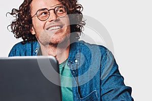Closeup studio shot of a young smiling man with curly hair sitting at table working at laptop and dreaming. Businessman wearing