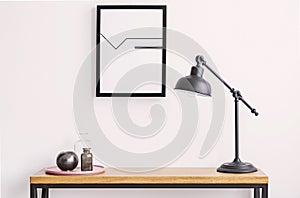 Closeup of table with lamp and plate with glass bottles, poster in black frame on white wall
