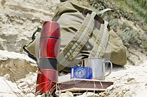 Closeup of thermos, notebook,compass and metall mug on the rock, backpack as a background