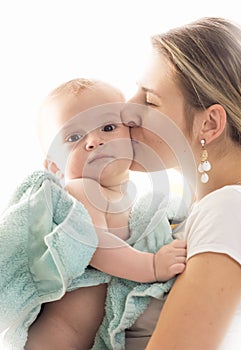 Closeup toned portrait of young loving mother kissing her baby son covered in blue towel after having bath