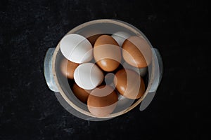 Closeup top view of handmade ceramic bowl of fresh organic brown and white eggs on dark moody background.