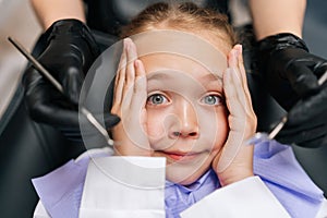 Closeup top view portrait of cute afraid child girl holding head from fear with hand, sitting on dentist chair. Close-up