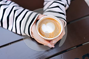 Closeup top view of unrecognizable young woman holding hot cup of coffee with heart shape. Close up cropped shot of