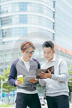 Closeup of two smiling business men using tablet computer and walking with office building in background - Image