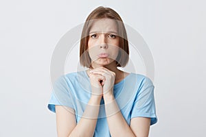 Closeup of upset unhappy young woman in blue t shirt biting her lip and looks abused isolated over white wall Keeps hands folded