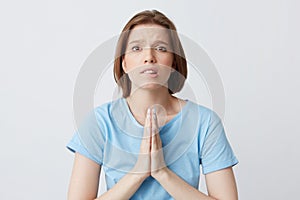 Closeup of upset worried young woman in blue t shirt keeps hands folded in praying position and biting her lip isolated over white