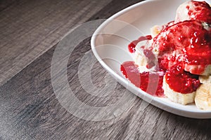 Closeup Vanilla ice cream poured with strawberry or raspberry jam with sliced banana slices on a white plate on a dark