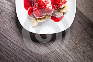 Closeup Vanilla ice cream poured with strawberry or raspberry jam with sliced banana slices on a white plate on a dark