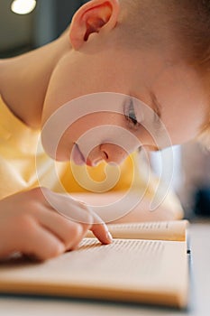 Closeup vertical shot of cute pupil schoolboy reading paper book following lines with finger along page sitting at desk