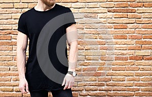 Closeup view of bearded muscular man wearing black tshirt and jeans posing outside. Empty brown grunge brick wall on the