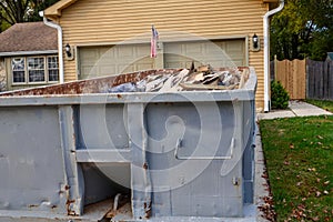 Closeup view of a blue dumpster filled with construction debris in the driveway of a yellow house in front of the garage doors