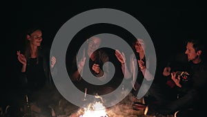 Closeup view of the bonfire late at night. Young people sitting by the fire in the evening, playing guitar. Cheerful