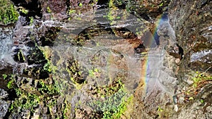 Closeup view of cascade with water falling from stone rock covered by plants and small rainbow