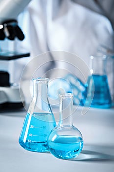 Closeup view of scientist pouring light blue liquid from test tube into flask and laboratory glassware on table, selective focus