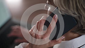 Closeup view from shoulder of businesswoman talking on smartphone siting at office desk working typing on laptop