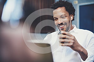 Closeup view of Smiling african man using smartphone to listen to music while sitting on the bench at sunny street