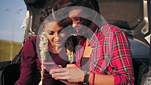 Closeup view of two young attractive girlfriends sitting inside of the open car trunk in the parking by the shopping