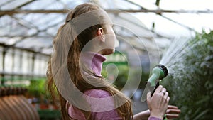 Closeup view of young attractive female gardener in uniform watering plants with garden hose in greenhouse. Slowmotion