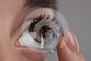 Closeup view of young woman putting contact lens in her eye against grey background