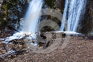 Closeup of waterfall. Colored rocks in cliff. Wood and sand in foreground.