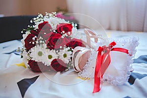Closeup of wedding rings on a pillow with a red ribbon on a bed with a flower bouquet on it