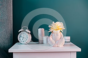 Closeup white bedside table with clock, female body-shaped vase with rose flower and candles on turquoise wall