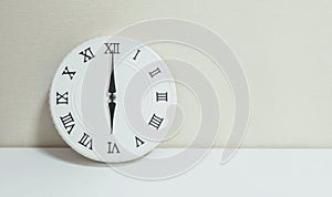 Closeup white clock for decorate in 6 o`clock on white wood desk and cream wallpaper textured background with copy space