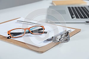 Closeup of white desktop with laptop, glasses, coffee cup, notepads and other items on blurry city background