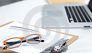 Closeup of white desktop with laptop, glasses, coffee cup, notepads and other items on blurry city background