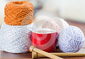 Closeup white, orange yarn ball with knitting needles and scarf in progress lying on desk, coffee mug sitting next to it