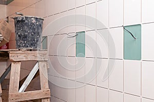 Closeup white tile on the wall of drywall without grout and electrical wires, scaffold, bucket with adhesive. Concept of repair in