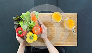 Closeup woman hand holding glass bowl with variety of vegetable on wooden chop board. Bell pepper, tomato, carrot and lettuce.