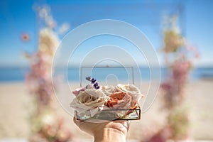 Closeup woman hands hold glass box for wedding rings decorated with fresh rose flowers and banch of lavender at blurred coast