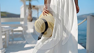 Closeup of woman in long white dress holding summer straw hat and walking on white wooden pier