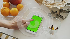 Closeup of woman`s hand browsing smartphone with green screen on kitchen table at home