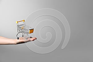 Closeup of a woman`s hand holding a small metal empty shopping cart in the palm of her hand on gray background. The concept of