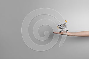 Closeup of a woman`s hand holding a small metal empty shopping cart in the palm of her hand on gray background. The concept of