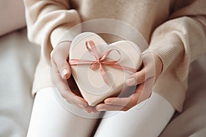 Closeup woman& x27;s hands holding Valentine& x27;s gift box in shape of heart, sitting on sofa at home