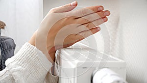 Closeup of woman warming her hands at heating radiator in house. Concept of energy crisis, high bills, broken heating system,