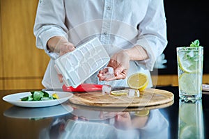 Closeup of a woman& x27;s hands preparing cold cocktail with lemon, mint leaves and ice