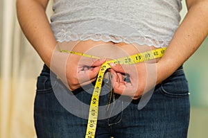 Closeup womans stomach with shirt lifted up, wearing jeans, measuring waistline using measure band, weightloss concept photo