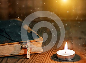 Closeup of wooden Christian cross on bible, burning candle on the old table.