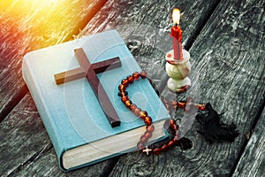 Closeup of wooden Christian cross on bible, burning candle and prayer beads on the old table.