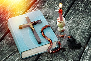 Closeup of wooden Christian cross on bible, burning candle and prayer beads on the old table.